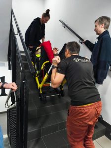 Trzy osoby na klatce schodowej przenoszą manekina na krześle ewakuacyjnym