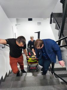 Trzy osoby na klatce schodowej. Dwoje wciąga manekina na krześle ewakuacyjnym w górę