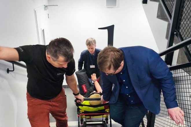 Trzy osoby na klatce schodowej. Dwoje wciąga manekina na krześle ewakuacyjnym w górę